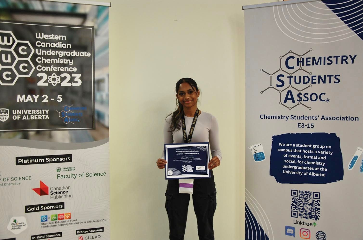 Chantell Fernandes standing between posters for the Western Canadian Undergraduate Chemistry Conference 2023 and the Chemistry Students Association.