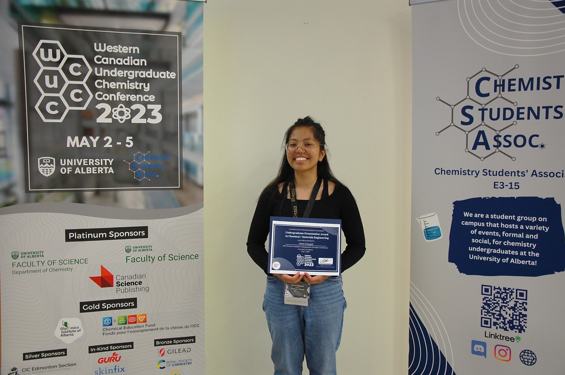 Maria Hangad standing between posters for the Western Canadian Undergraduate Chemistry Conference 2023 and the Chemistry Students Association.